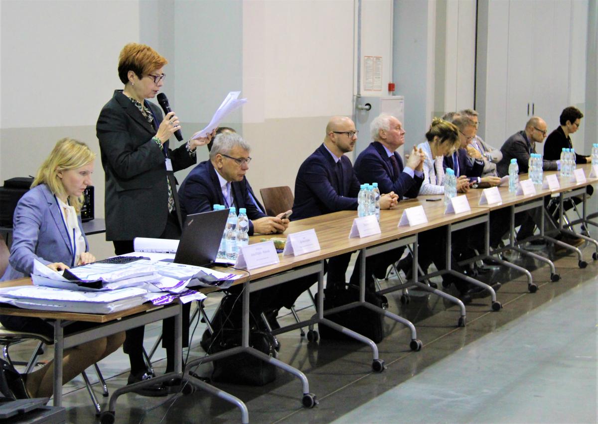 widok dużego prostokątnego stołu, przy którym znajduje się dwanaście osób; jedna z kobiet stoi trzymając w jednej ręce mikrofon, w drugiej kartkę papieru, pozostałe osoby siedzą