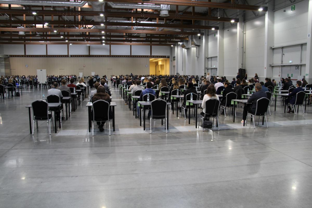 liczna grupa osób siedzących przy pojedynczych stolikach na dużej hali; z przodu, w środkowej części sali, w przejściu między stolikami stoi biała tablica ustawiona tyłem do obiektywu aparatu