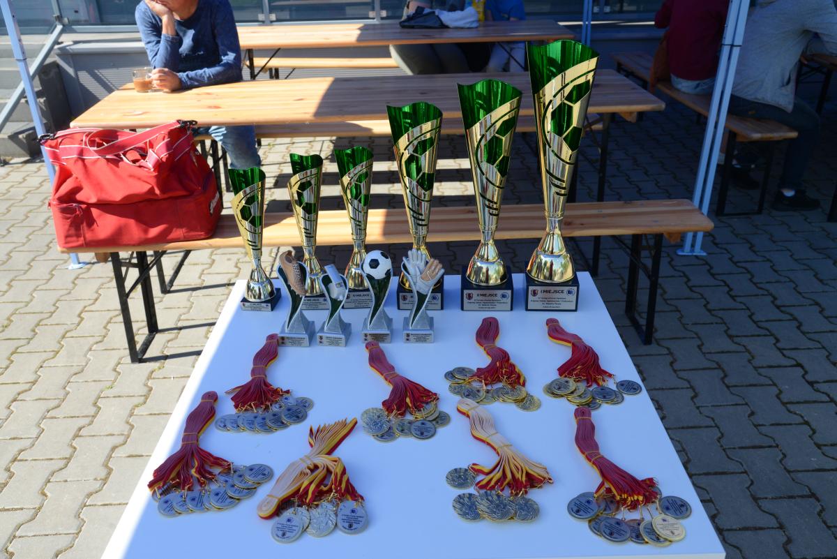 Zdjęcie nr:  1-DSC_3165_puchary i medale  przedstawia biały stolik na którym w trzech rzędach ułożone są nagrody dla piłkarzy takie jak: medale, statuetki, puchary. W pierwszym rzędzie na stoliki ułożone są okrągłe, w odcieniu złota medale z żółtoczerwoną wstążką, w drugim rzędzie  znajdują się cztery statuetki – pierwsza od lewej ma znaczek buta z przeznaczeniem dla króla strzelców, druga od lewej ma gwizdek z przeznaczeniem dla sędziego turnieju, trzecia ma znaczek piłki i jest przeznaczona dla najlepszego piłka a czwarta na znaczek dwóch rękawic bramkarskich z przeznaczeniem dla najlepszego bramkarza turnieju. W trzecim rzędzie znajduje się sześć zielonozłotych pucharów, różnej wielkości, ułożonych od najmniejszego do największego (od lewej strony). 