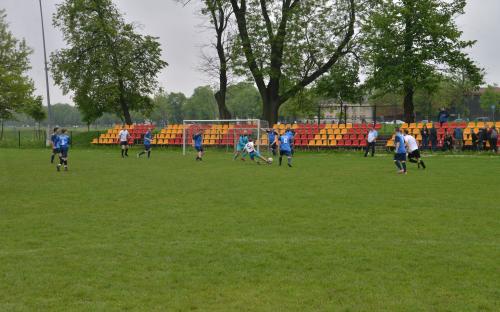 Zdjęcie nr 10: jedenastu mężczyzn na boisku piłkarskim biegnących za piłką w czasie meczu piłki nożnej, w tle bramka oraz czerwono żółte krzesła na trybunach