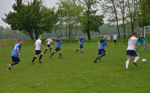 Zdjęcie nr 11: siedmiu mężczyzn na boisku piłkarskim biegnących za piłką w kierunku bramki, na której stoi bramkarz, w czasie meczu piłki nożnej, w oddali spacerująca osoba