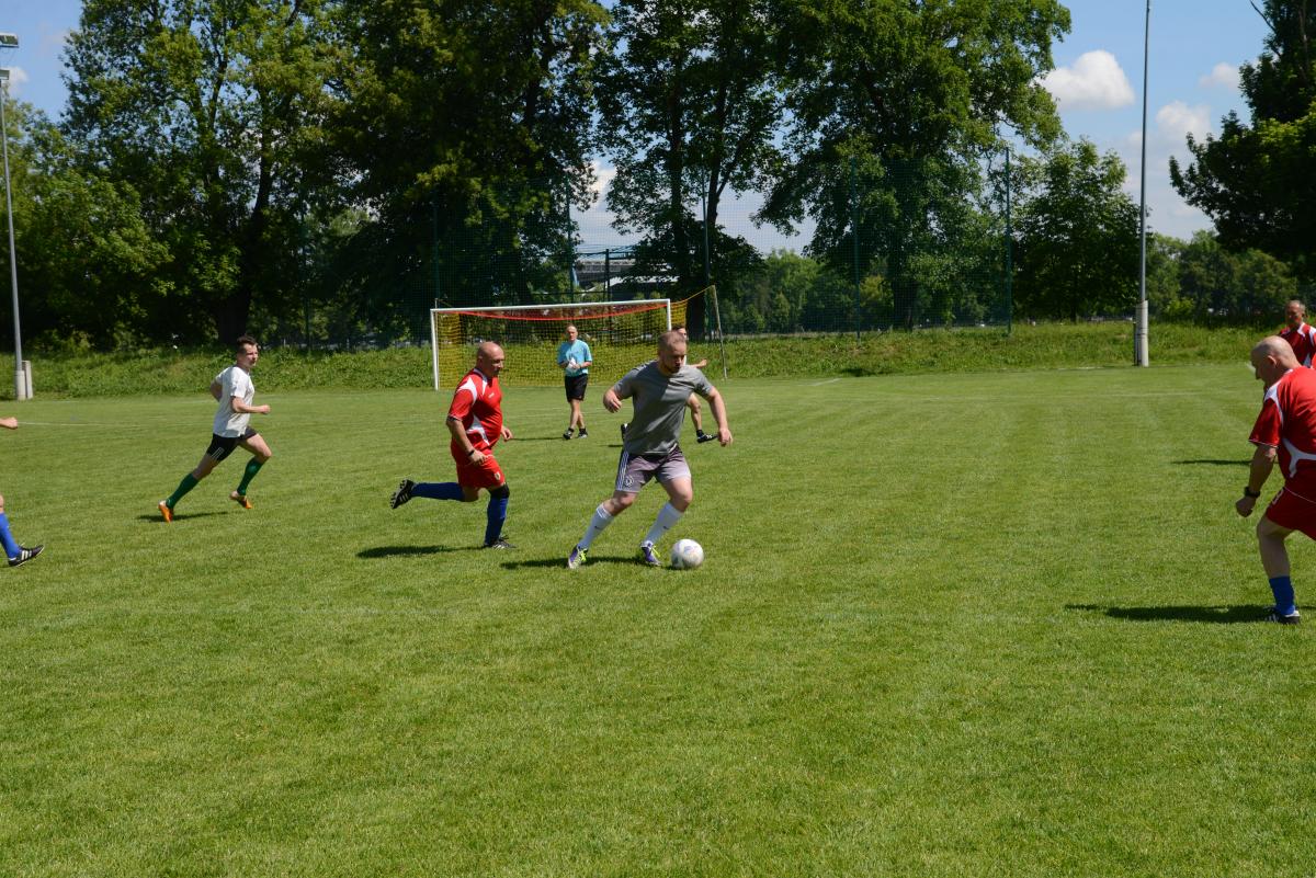 Zdjęcie nr: 12-DSC_3249 przedstawia odbywający się na murawie mecz piłkarski drużyn ubranych w jasnoczarne stroje oraz w stroje czerwone. Jeden z mężczyzn ma piłkę przy nodze.