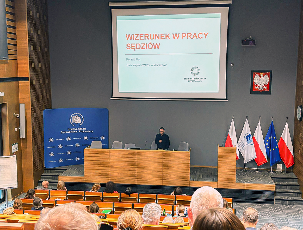 Sala konferencyjna Krajowej Szkoły Sądownictwa i Prokuratury, wykład o wizerunku sędziów prowadzony przez prelegenta stojącego przy mównicy, wyświetlony duży ekran z tytułem prezentacji, na scenie flagi Polski, Unii Europejskiej i KSSiP, siedzący uczestnicy.