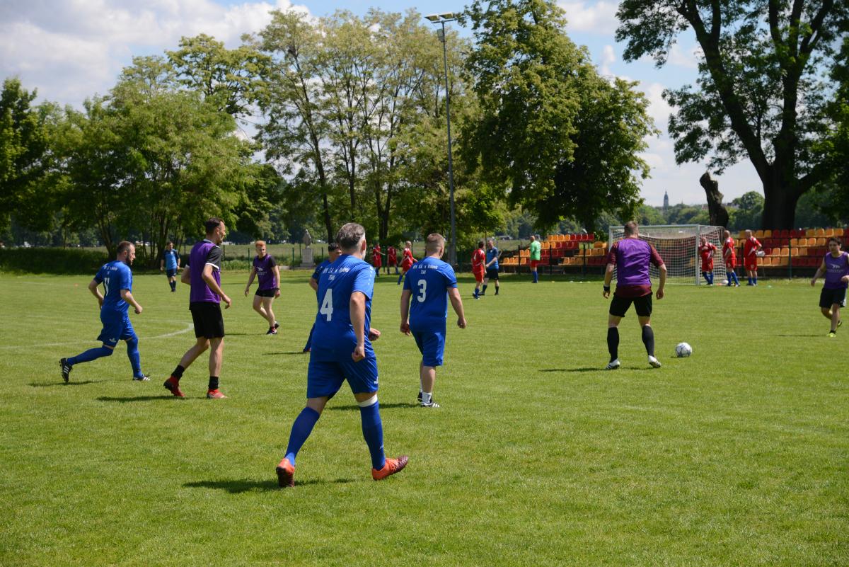 Zdjęcie nr: 15-DSC_3272 przedstawia odbywający się na murawie mecz piłkarski drużyn ubranych w niebieskie  stroje oraz w stroje fioletowoczarne.