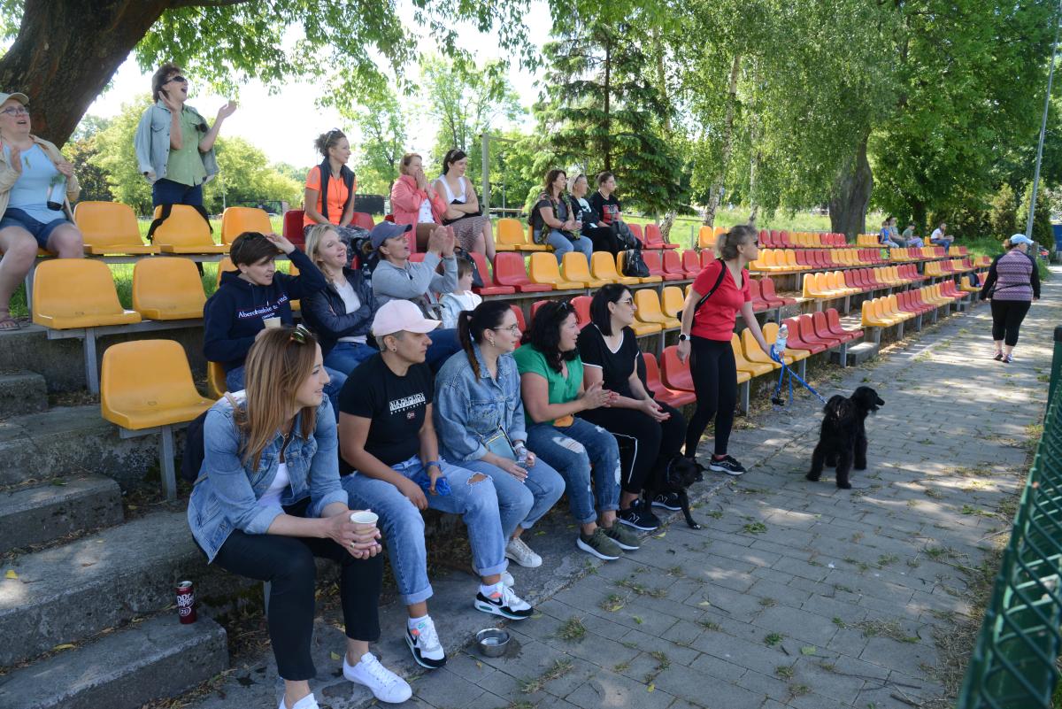 Zdjęcie nr: 18-DSC_3262 przedstawia trybuny z kibicami płci żeńskiej, siedzącymi na złotoczerwonych krzesełkach. Cześć kibiców ogląda, cześć dopinguje. W pierwszym rzędzie meczowi przygląda się także pies koloru czarnego z właścicielką.