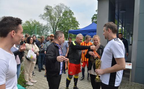 Zdjęcie nr 18: kobieta z medalami w rękach, obok niej trzech mężczyzn, jeden z ręką wyciągniętą w kierunku medali, drugi z pucharem w ręce, przygląda im się grupa osób, w tle widać niebieski dach zadaszenia namiotowego