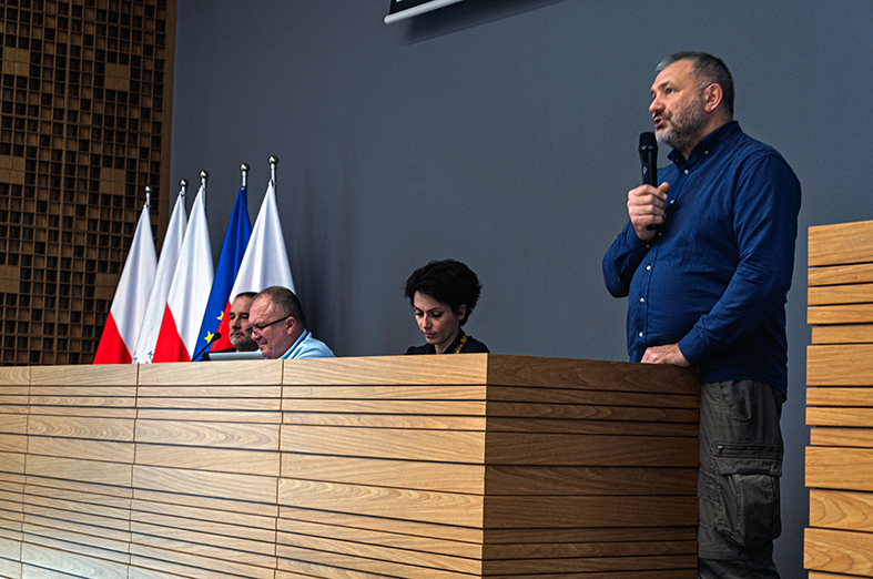 Na zdjęciu widoczna jest scena, na której znajdują się trzy osoby. Jedna z nich stoi i trzyma mikrofon, przemawiając do publiczności. Pozostałe dwie osoby siedzą za drewnianym stołem. W tle znajdują się flagi – dwie polskie oraz flaga Unii Europejskiej. Pomieszczenie ma nowoczesny wystrój, z drewnianymi elementami na ścianie i minimalistycznym designem.