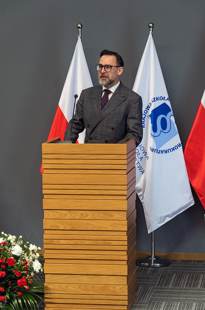 Na zdjęciu widać mężczyznę w okularach i garniturze stojącego za drewnianym podium. Za nim znajdują się dwie flagi, jedna polska i jedna z logiem Krajowej Szkoły Sądownictwa i Prokuratury. Po lewej stronie podium widać bukiet kwiatów.