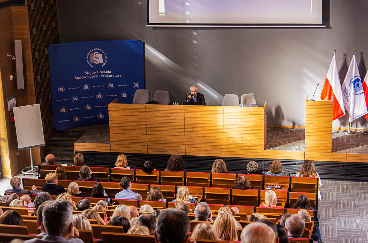 Widok sali konferencyjnej z uczestnikami siedzącymi w ławkach, z przodu znajdują się dwie osoby za stołem prezydialnym, a trzecia stoi za mównicą. Po prawej stronie stoją flagi Polski, flaga szkolna oraz flaga Unii Europejskiej.