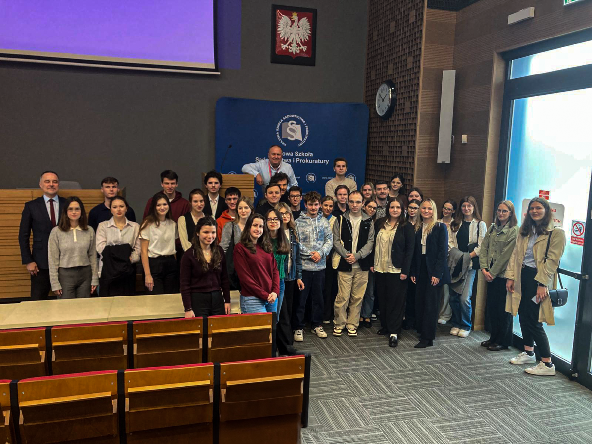 Na zdjęciu widzę grupę ludzi, studentów wraz z wykładowcą i osobą prowadzącą, pozujących do zdjęcia. W tle widoczny jest godło Polski, a także logo Krajowej Szkoły Sądownictwa i Prokuratury. Zdjęcie zostało zrobione w auli, o czym świadczą rzędy krzeseł widoczne w dolnej części kadru.