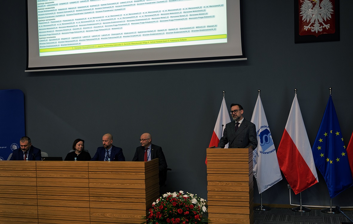 Na zdjęciu widać salę konferencyjną lub miejsce oficjalnego wydarzenia. Na pierwszym planie znajduje się mównica, przy której stoi mężczyzna w garniturze. Po prawej stronie widoczne są flagi Polski, Unii Europejskiej oraz organizacji związanej z wydarzeniem. Na lewo od mównicy znajduje się stół, przy którym siedzi kilka osób, również ubranych formalnie. Na ścianie wisi godło Polski, a na ekranie wyświetlany jest slajd z listą nazwisk i informacjami, które mogą być związane z programem lub uczestnikami wydarzenia. Dekorację uzupełniają kwiaty ustawione przed stołem.