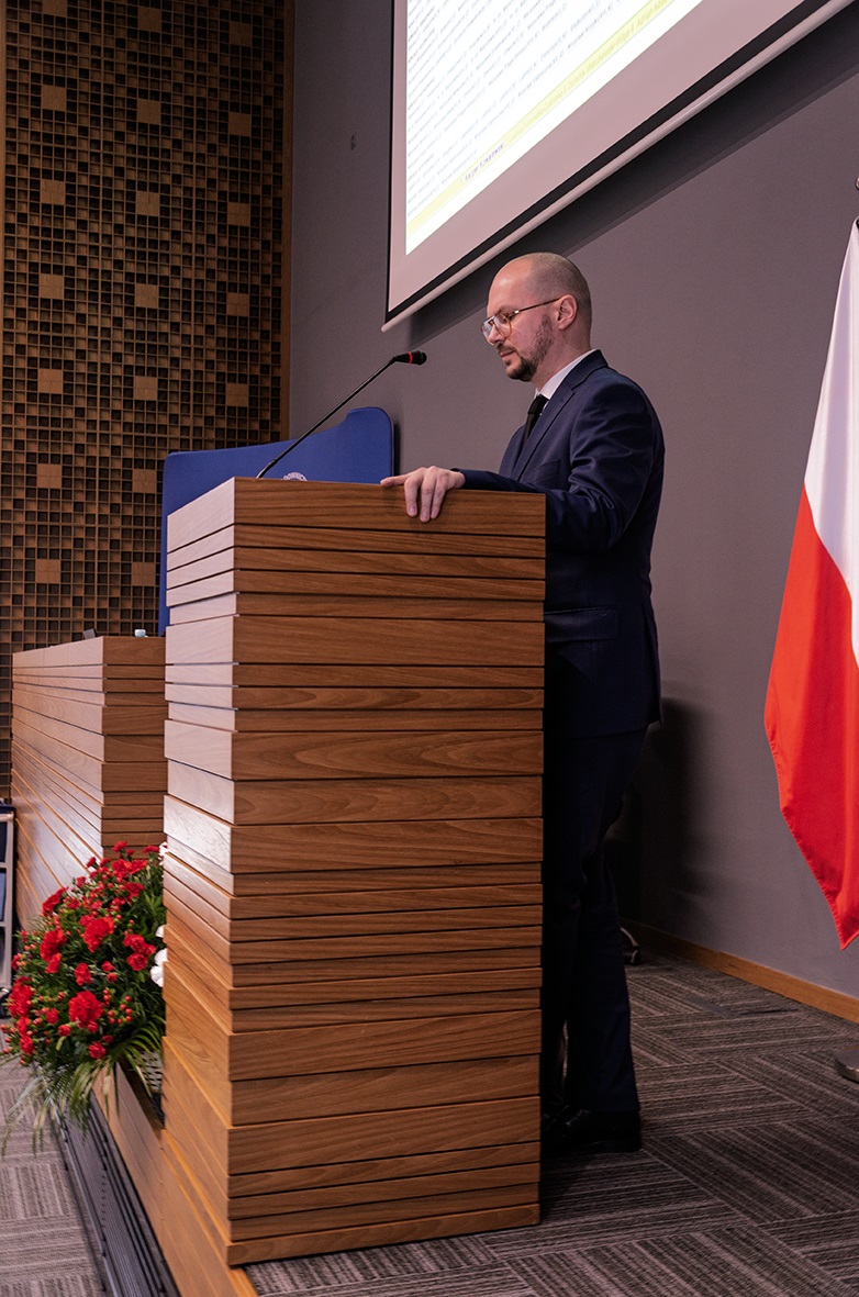 Na zdjęciu widać mężczyznę w garniturze stojącego za drewnianym podium z mikrofonem. Za nim znajduje się ekran projekcyjny i flaga Polski. W tle widać ścianę z dekoracyjnym wzorem. Z boku podium stoi kompozycja kwiatowa z czerwonych i białych kwiatów.