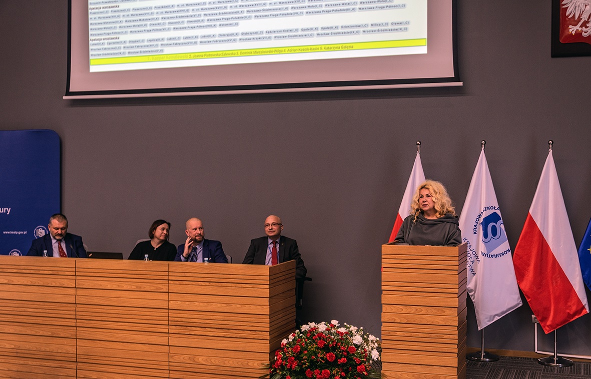 Na zdjęciu widzimy osobę przemawiającą przy drewnianej mównicy w sali konferencyjnej. W tle znajduje się ekran z prezentacją, a po prawej stronie widoczna jest flaga Polski. Na dole mównicy umieszczono dekorację z czerwonych i białych kwiatów.