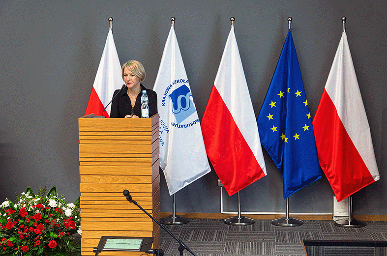 Na zdjęciu znajduje się osoba stojąca za drewnianą mównicą, przemawiająca na tle flag. Po lewej stronie i po prawej stronie widoczne są dwie polskie flagi z biało-czerwonymi pasami. Między nimi znajdują się flaga Unii Europejskiej z niebieskim tłem i żółtymi gwiazdami oraz biała flaga z logo Krajowej Szkoły Sądownictwa i Prokuratury. Na mównicy stoi butelka wody. Po lewej stronie zdjęcia widoczny jest fragment czerwono-białych kwiatów, które stanowią dekorację sali. Całość znajduje się w pomieszczeniu o szarym tle, które nadaje formalny charakter wydarzeniu.