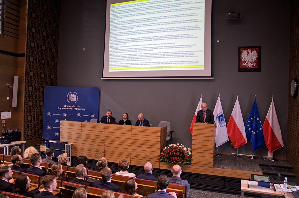 Na zdjęciu widać salę konferencyjną lub wykładową, w której odbywa się oficjalne wydarzenie. Na pierwszym planie znajdują się uczestnicy siedzący w rzędach, a na podium widoczne są osoby przemawiające lub prowadzące spotkanie. Za mównicą znajdują się flagi Polski oraz Unii Europejskiej, a także tablica z logo instytucji "Krajowa Szkoła Sądownictwa i Prokuratury". Na ścianie wisi godło Polski, a na ekranie wyświetlona jest prezentacja z tekstem. Dekorację podium uzupełnia bukiet czerwonych kwiatów.