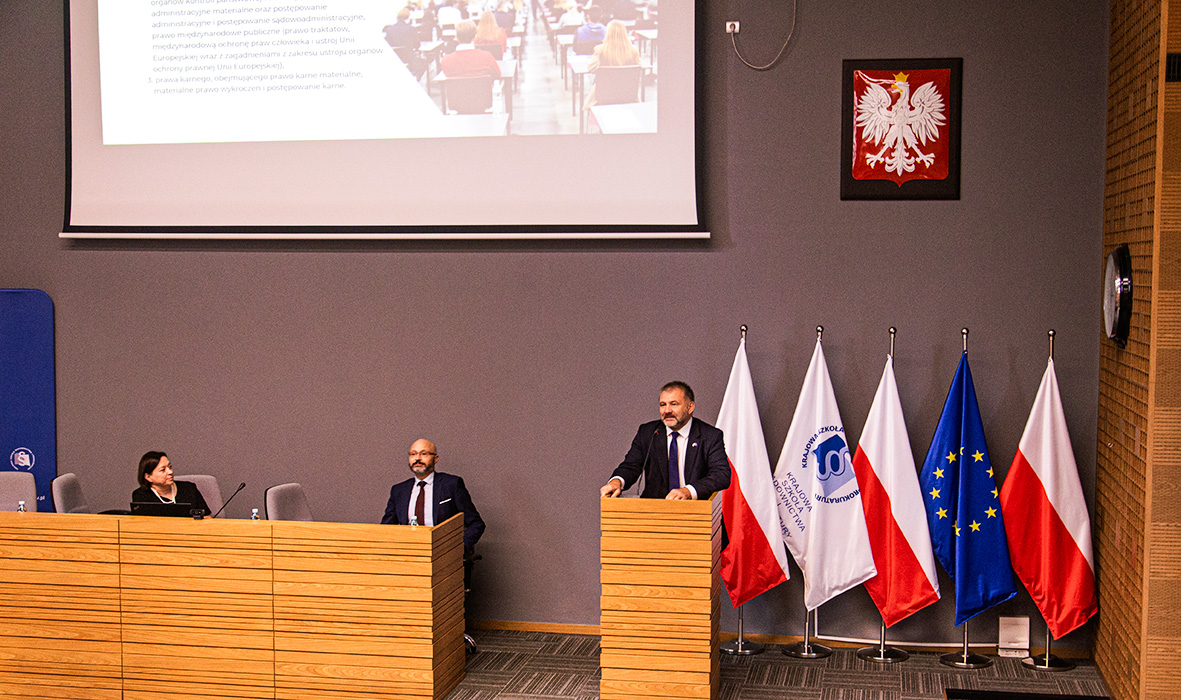 Ujęcie w pełni zajętej sali wykładowej z osobami siedzącymi na drewnianych ławkach, skupionymi na prelegentach i prezentacji wyświetlanej na ekranie. Po prawej stronie widoczne są stojące flagi Polski, Unii Europejskiej i szkolna flaga KSSiP.