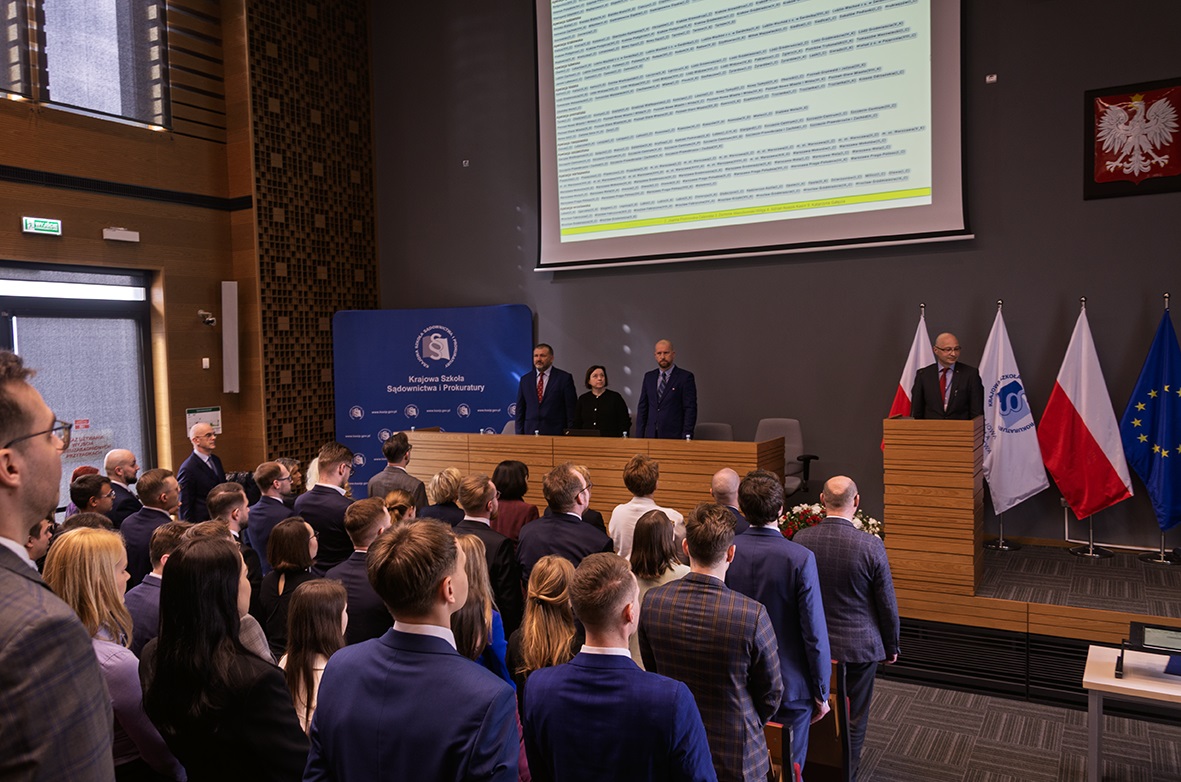 Na zdjęciu widać salę konferencyjną lub audytoryjną, w której zgromadziła się grupa ludzi, prawdopodobnie podczas jakiegoś wydarzenia. W przedniej części sali znajduje się scena z mównicą, za którą stoją osoby przemawiające. W tle widoczne są flagi Polski i Unii Europejskiej oraz baner Krajowej Szkoły Sądownictwa i Prokuratury. Na ścianie wyświetlany jest ekran projekcyjny z tekstem. Większość osób na widowni jest ubrana w garnitury, co sugeruje formalny charakter wydarzenia.