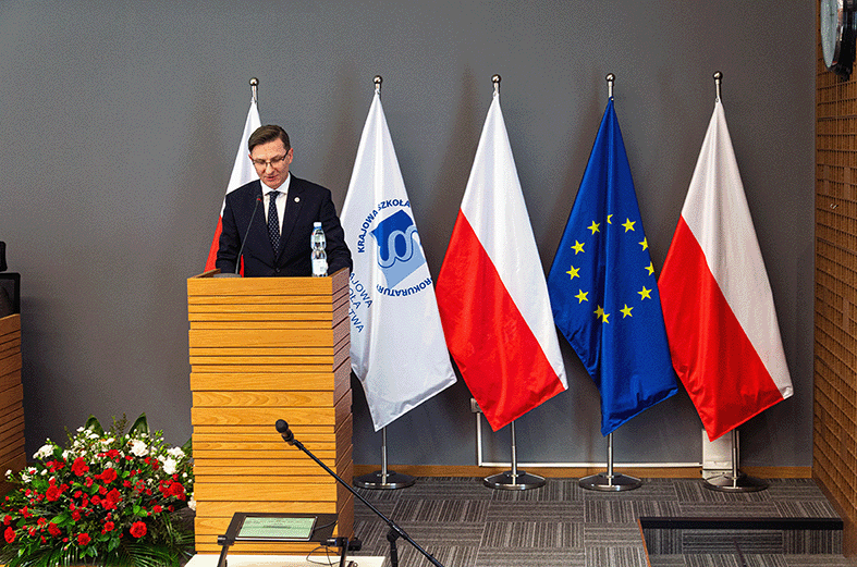 Na zdjęciu widać mężczyznę w okularach i ciemnym garniturze z czerwoną przypinką w klapie, który stoi za drewnianą mównicą. Za nim znajdują się cztery flagi: dwie polskie z białym i czerwonym pasem, flaga Unii Europejskiej z niebieskim tłem i żółtymi gwiazdami oraz biała flaga z logo Krajowej Szkoły Sądownictwa i Prokuratury. Po lewej stronie u dołu znajdują się biało-czerwone kwiaty. Na mównicy widać mikrofon. Ściana w tle jest szara.