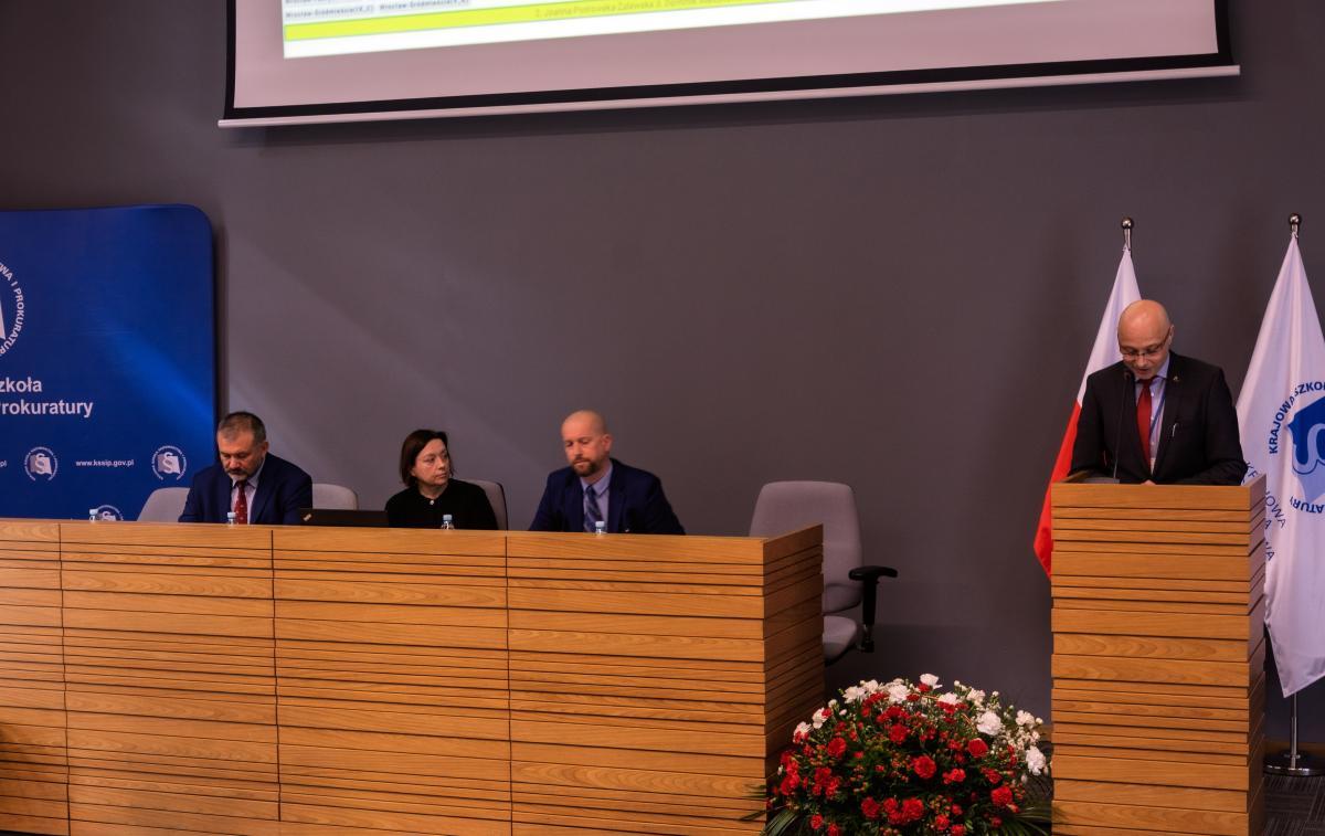Na zdjęciu widać salę konferencyjną z kilkoma osobami. Na pierwszym planie znajduje się drewniana mównica, za którą stoi mężczyzna w garniturze i czerwonym krawacie, prawdopodobnie wygłaszający przemówienie. Za nim widoczne są dwie flagi. Po lewej stronie mównicy widać stół, za którym siedzą trzy osoby: mężczyzna, kobieta i kolejny mężczyzna. W tle widać szary mur oraz ekran projekcyjny. Przed stołem znajduje się dekoracja z czerwonych i białych kwiatów.