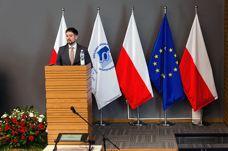 Na zdjęciu widoczna jest kobieta stojąca za drewnianą mównicą. Jest ubrana w ciemną bluzkę. Za nią znajdują się cztery flagi: dwie polskie z białym i czerwonym pasem, flaga Unii Europejskiej z niebieskim tłem i żółtymi gwiazdami oraz biała flaga z logo Krajowej Szkoły Sądownictwa i Prokuratury. Po lewej stronie zdjęcia, na dole, znajdują się biało-czerwone kwiaty.