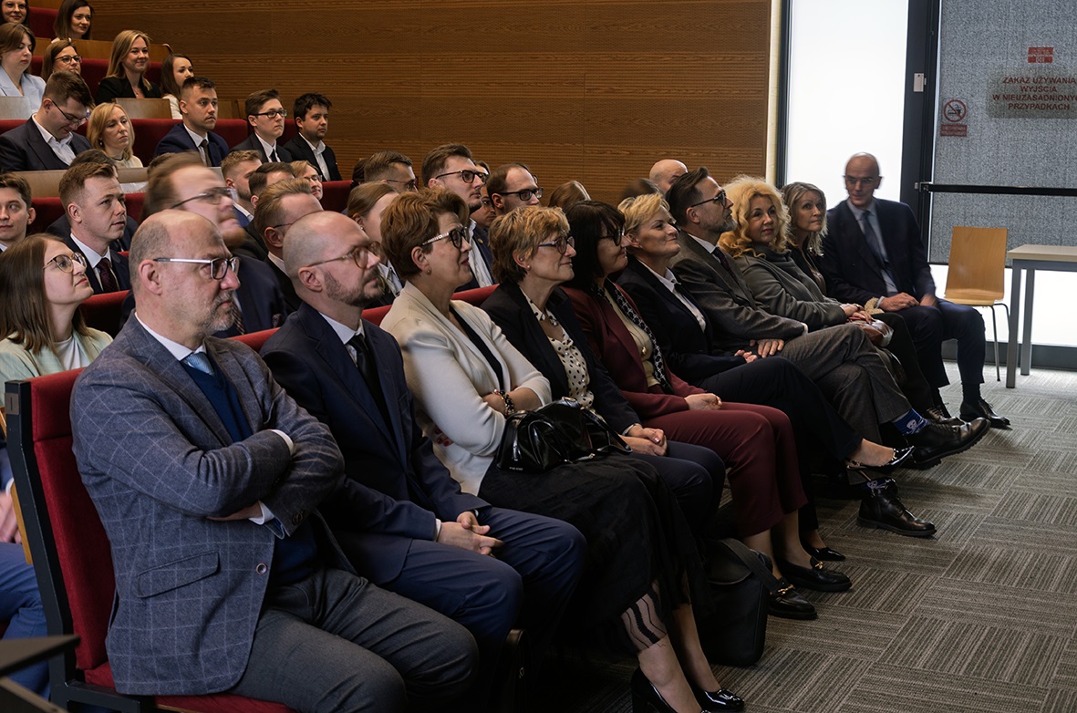 Na zdjęciu widać grupę ludzi siedzących w rzędach, prawdopodobnie podczas konferencji lub prezentacji. Większość osób jest ubrana formalnie. W tle widać drewniane panele i drzwi.