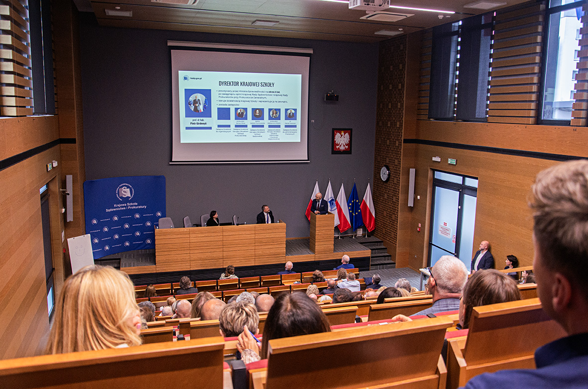 Ujęcie szerokie przedstawiające całą salę wykładową z rzędem siedzących osób, prelegentem za mównicą oraz widocznymi flagami Polski, KSSiP i Unii Europejskiej.