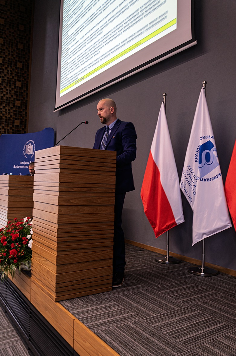 Na zdjęciu widać mężczyznę w garniturze stojącego za drewnianym podium z mikrofonem. Za nim znajdują się trzy flagi, w tym flaga Polski, oraz ekran projekcyjny. Po lewej stronie podium widać dekorację z czerwonych i białych kwiatów.