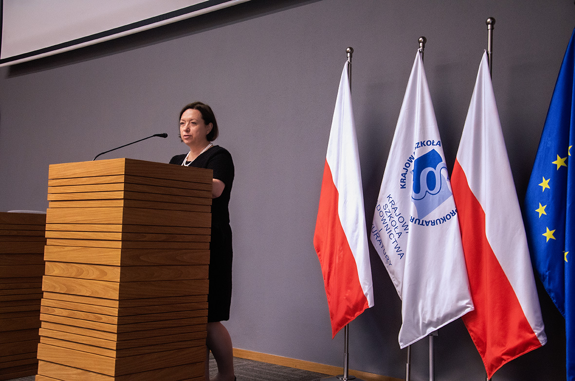 •	Ujęcie z góry na salę konferencyjną z uczestnikami zwróconymi w kierunku prezentacji wyświetlanej na ekranie. Po prawej stronie stoją flagi Polski i Unii Europejskiej, przy mównicy przemawia prelegent.
