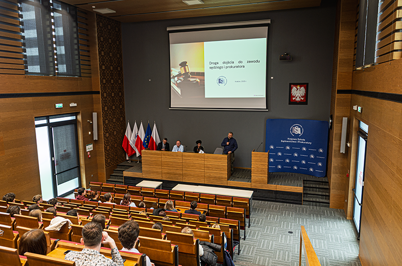Na zdjęciu widać salę wykładową lub konferencyjną, w której odbywa się prezentacja. Na pierwszym planie znajdują się uczestnicy siedzący w rzędach na drewnianych ławkach. Na środku zdjęcia widoczna jest katedra, gdzie siedzi kilka osób prowadzących spotkanie. Na ekranie wyświetlany jest slajd z tytułem "Droga dojścia do zawodu sędziego i prokuratora", co sugeruje, że tematem wydarzenia jest kariera w zawodach prawniczych. W tle znajdują się flagi Polski oraz Unii Europejskiej, a także godło Polski na ścianie, co wskazuje na oficjalny charakter spotkania. Po prawej stronie widoczna jest niebieska ścianka z logo instytucji, prawdopodobnie związanej z wymiarem sprawiedliwości lub edukacją prawniczą. Cała sala ma nowoczesny wystrój, z drewnianymi elementami i dużymi oknami po lewej stronie.