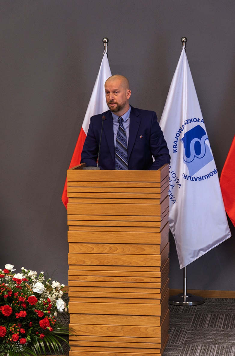Na zdjęciu widać mężczyznę stojącego za drewnianym podium. Mężczyzna ma krótkie włosy i brodę, ubrany jest w ciemny garnitur, koszulę i krawat. Za nim widoczne są dwie flagi, prawdopodobnie Polski i Krajowej Szkoły Sądownictwa i Prokuratury. Po lewej stronie podium stoi bukiet czerwonych i białych kwiatów. Tło jest szare.