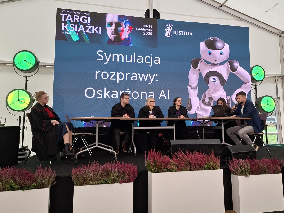 Na scenie siedzi pięć osób – cztery w togach sędziowskich oraz jedna w marynarce – uczestniczące w symulowanej rozprawie sądowej pod tytułem „Oskarżona AI”. W tle widać duży ekran z tytułem wydarzenia oraz grafiką humanoidalnego robota. Na pierwszym planie ustawione są donice z wrzosami, a po bokach sceny stoją zielone reflektory. Wydarzenie odbywa się w ramach 28. Międzynarodowych Targów Książki w Krakowie i pokazuje edukacyjne wykorzystanie sztucznej inteligencji w formie interaktywnego spektaklu.