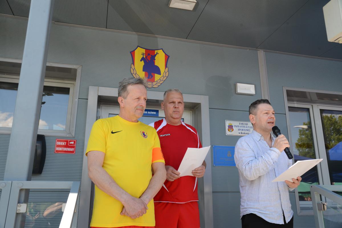 Zdjęcie nr: 1a-DSC_3198 przedstawia trzech mężczyzn na tle szarego budynku, z tyłu za ich głowami znajduje się herb Zwierzynieckiego Klubu Sportowego; mężczyzna od lewej jest ubrany w żółtą koszulkę sportową z logo Zwierzynieckiego Klubu Sportowego i pomarańczową opaską na lewym ramieniu, mężczyzna w środku jest ubrany w strój sportowy koloru czerwonego z białymi cienkimi paskami, w rękach trzyma kartkę, trzeci mężczyzna od lewej ma błękitną koszulę i czarne spodnie, w prawej ręce trzyma mikrofon, ponieważ przemawia – natomiast w lewej ręce ma kartkę.