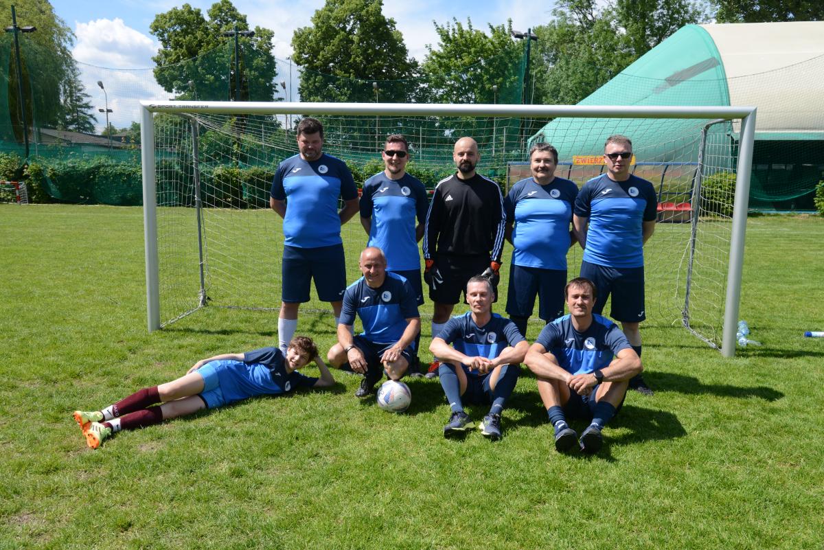 Zdjęcie nr : 2-DSC_3385 przedstawia skład męskiej drużyny piłkarskiej, składającej się z dziewięciu zawodników, na tle bramki piłkarskiej, na murawie, ośmiu z nich ubranych jest w granatowoczarne stroje a jeden w strój bramkarski koloru czarnego. Pięciu mężczyzn stoi, pozostali spoczywają na trawie (jeden leży, jeden kuca, dwóch siedzi na trawie).