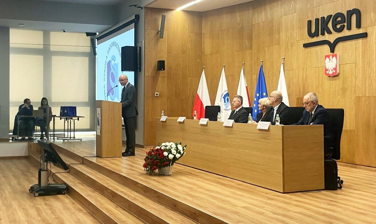 Na zdjęciu widać salę konferencyjną, gdzie mężczyzna w garniturze stoi za mównicą i wygłasza przemówienie. Za nim, przy stole prezydialnym, siedzą cztery osoby, a w tle widać flagi. Na pierwszym planie, obok mównicy, stoi kosz z czerwonymi i białymi kwiatami.