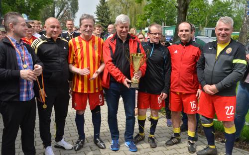 Zdjęcie nr 22: siedmiu mężczyzn, jeden z nich ma medal na szyi oraz trzyma medal w ręce, mężczyzna w środku grupy trzyma puchar, w tle grupa obserwatorów