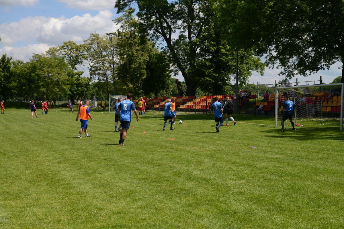Zdjęcie nr: 23-DSC_3339 przedstawia odbywający się na murawie mecz piłkarski drużyn ubranych w granatowoczarne stroje oraz w stroje niebieskie. Zawodnicy w niebieskim stroju piłkarskim są dodatkowo ubrani w pomarańczową koszulkę na ramiączkach. Zdjęcie przedstawia również zawodnika w bramce a w tle znajdują się  trybuny.