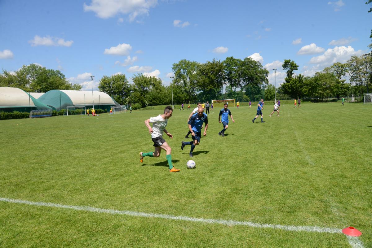 Zdjęcie nr: 25-DSC_3372 przedstawia odbywający się na murawie mecz piłkarski drużyn ubranych w jasnoczarne stroje oraz w stroje granatowe. Na pierwszym planie dwóch zawodników kopiących piłkę. W tle pozostali zawodnicy.