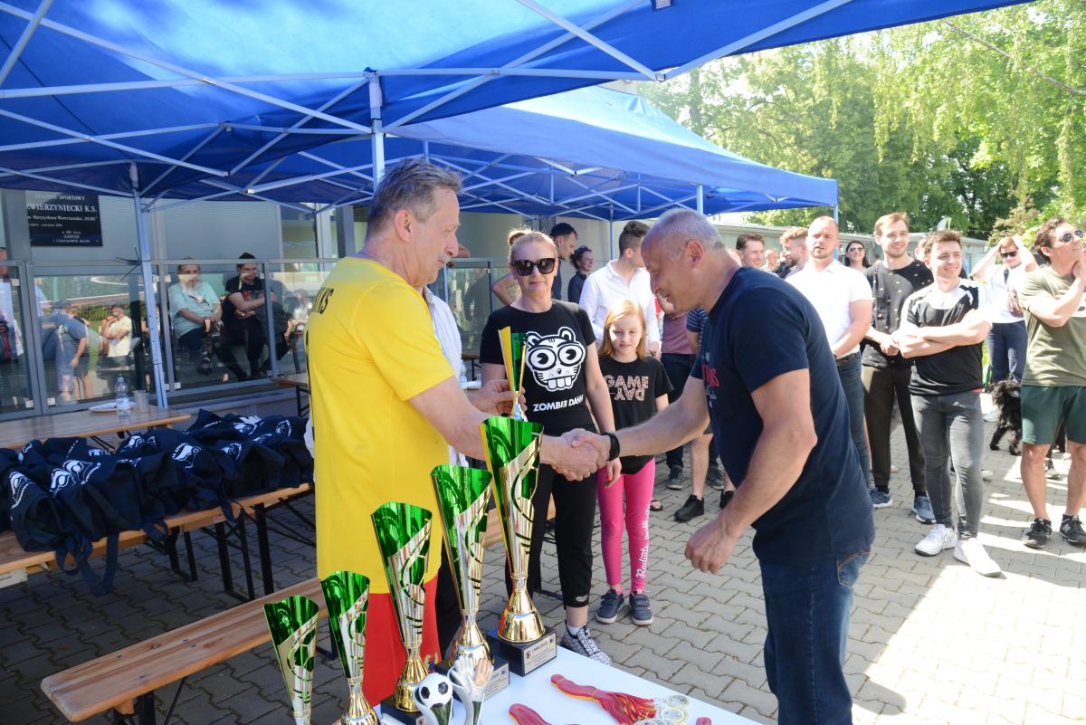 Zdjęcie nr: 26-DSC_3402 na pierwszym planie przedstawia wręczanie pucharu. Jeden z mężczyzn w lewej ręce trzyma puchar a drugą rękę podaje drugiemu mężczyźnie. Tuż obok stoi kobieta a za nią dziewczynka w wieku szkolnym. W pobliżu jest również stolik, na którym  znajdują puchary, statuetki i medale. W dalszym planie zdjęcie przedstawia mężczyzn-zawodników biorących udział w meczach oraz kibiców. Na zdjęciu znajdują się również stolik z nagrodami dla piłkarzy w bawełnianych torbach w kolorze granatowym z białym logo.