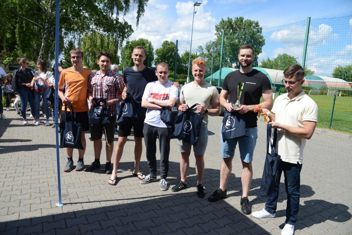 Zdjęcie nr: 28-DSC_3439 przedstawia siedmiu mężczyzn trzymających w rękach bawełniane torby w kolorze granatowym,  jeden z nich trzyma w ręce puchar, drugi medale. Na drugim planie -  zdjęcie przedstawia kibiców – kobiety; w tle znajduje się również boisko piłkarskie.