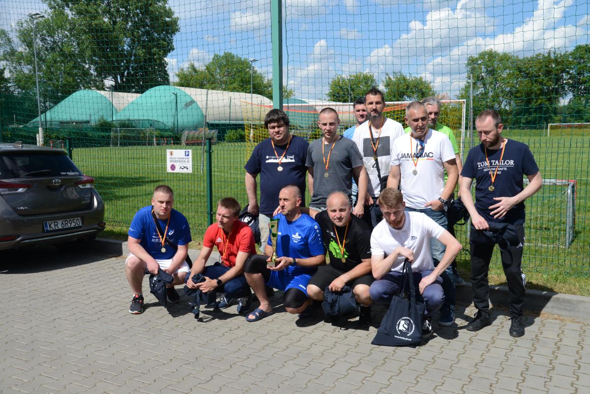 Zdjęcie nr: 29-DSC_3497 przedstawia dwunastu mężczyzn, jeden z nich trzyma puchar. Na szyjach mają powieszone medale, na żółtoczerwonych wstążkach, a w rękach trzymają granatowe torby w kolorze granatowym. Siedmiu mężczyzn stoi, pozostali kucają.  W tle znajduje się boisko piłkarskie.