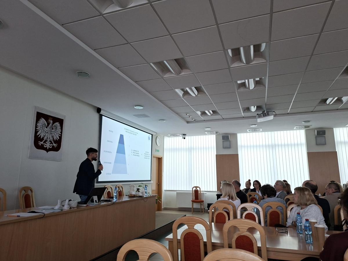 Sala konferencyjna z rzędami krzeseł i stołem prezydialnym. Prelegent stoi przy ekranie, na którym widoczny jest wykres w formie piramidy z napisami w języku polskim. Na sali siedzi grupa osób zwróconych w stronę prezentacji. Na ścianie wisi godło Polski.