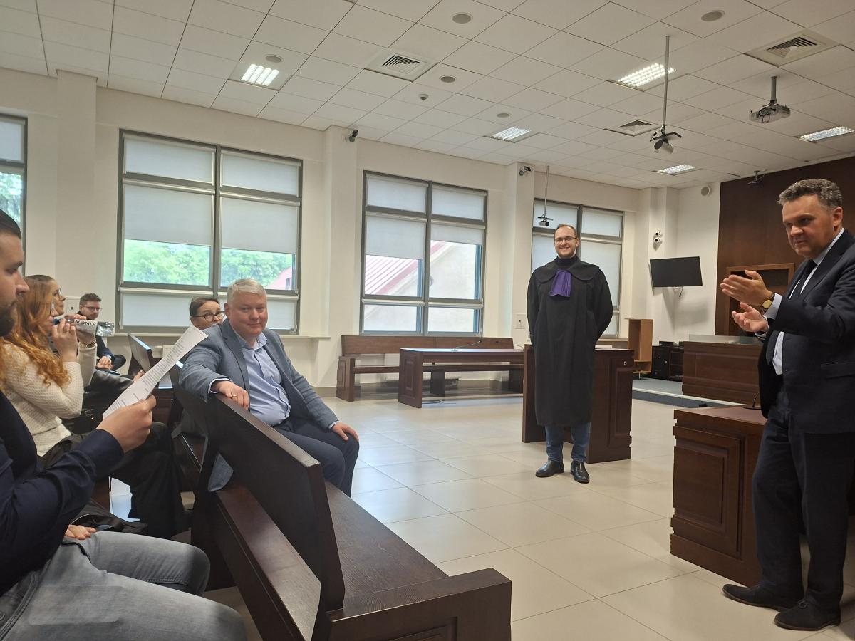 Wnętrze jasnej sali sądowej. Na pierwszym planie po lewej stronie siedzi grupa osób, niektórzy trzymają dokumenty. Po prawej stronie stoi mężczyzna w garniturze. Na środku sali stoi uśmiechnięty mężczyzna w todze z fioletowym żabotem. W tle widać ławy, okna z opuszczonymi roletami oraz sprzęt nagrywający na suficie.