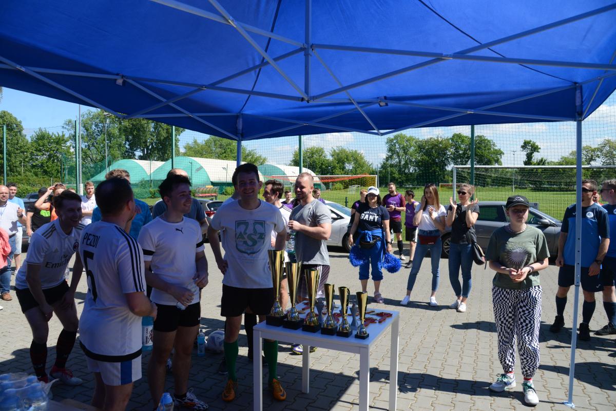 Zdjęcie nr: 2aDSC_3190 przedstawia zawodników jednej z drużyn, ubranych w jasne koszulki i czarne spodenki, obok nich stoi stolik na którym znajdują się puchary, medale i statuetki. Zdjęcie przedstawia również kibiców płci żeńskiej (na drugim planie). W tle znajduje się boisko piłkarskie. 