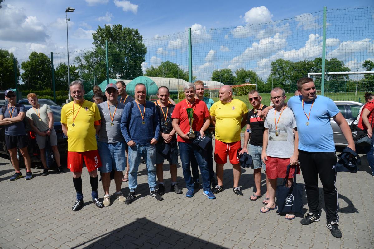 Zdjęcie nr: 30-DSC_3511 przedstawia jedenastu mężczyzn, a czterech z nich  trzyma w ręce bawełniane torby w kolorze granatowym. Na szyjach mają powieszone medale, na żółtoczerwonych wstążkach. Jeden z nich trzyma w ręce puchar.  Z boku stoi dwóch mężczyzn również z medalami na szyi żółtoczerwonymi wstążkami. W tle znajduje się boisko piłkarskie.