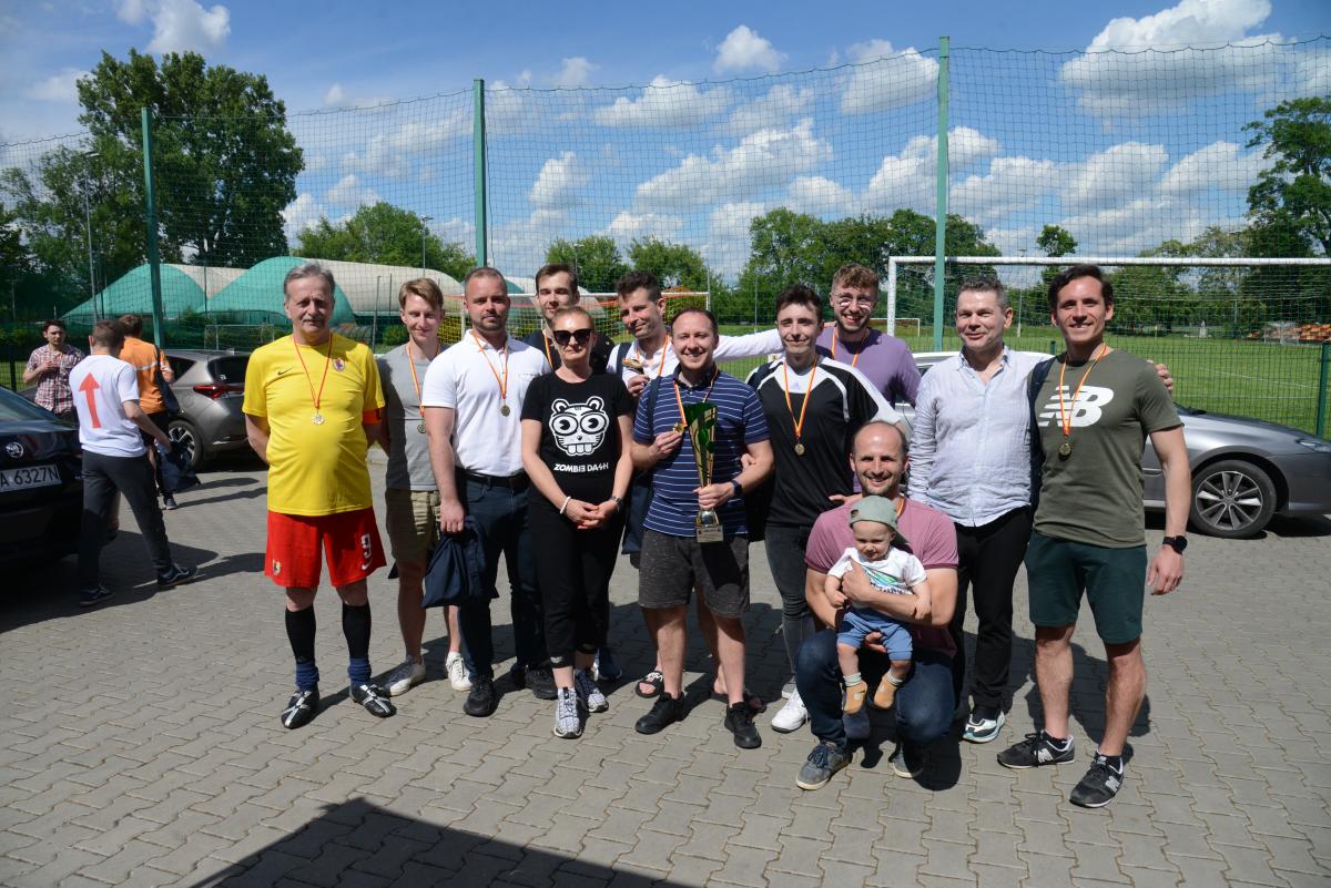Zdjęcie nr:  31-DSC_3529 przedstawia grupę osób: jedną kobietę, stojącą pomiędzy  jedenastoma mężczyznami z których jeden kuca, trzymając na rękach małe dziecko. Dziesięciu z nich na szyi ma powieszone medale, na żółtoczerwonych wstążkach. A dodatkowo jeden z nich trzyma w ręce puchar. W tle znajduje się boisko piłkarskie i inni trzej mężczyźni.