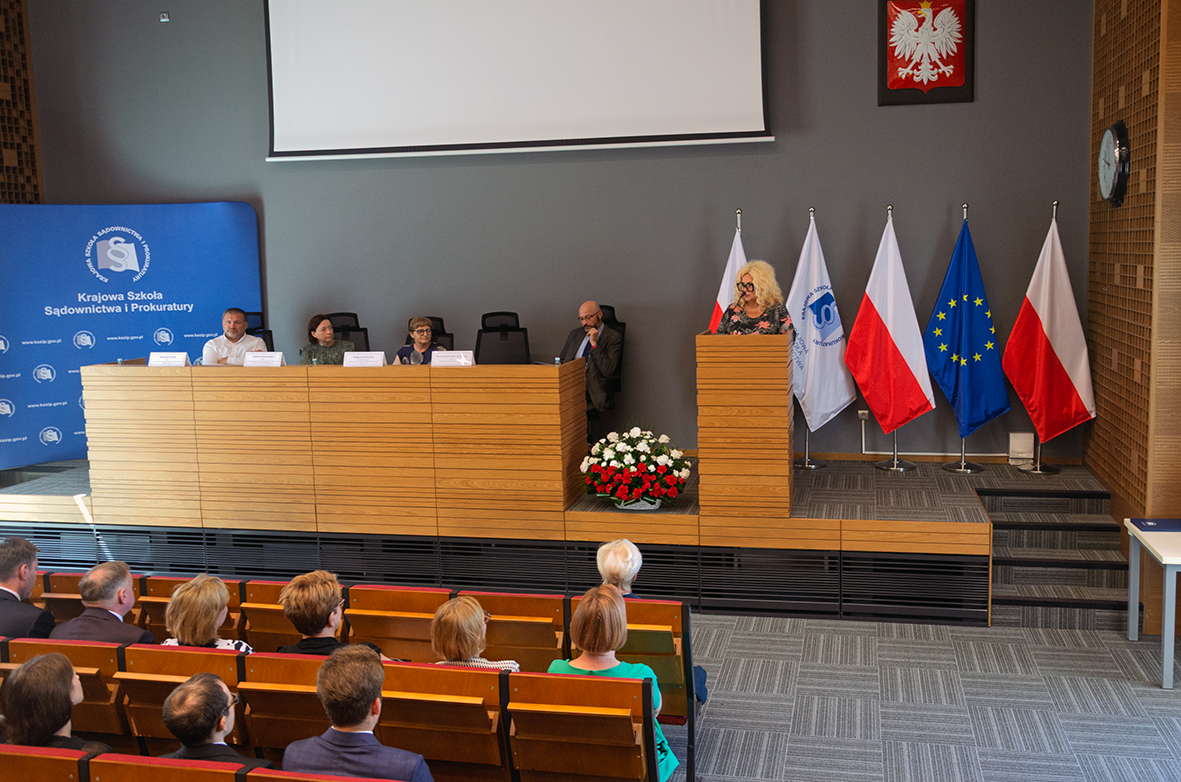Widok na scenę w sali konferencyjnej. Przy mównicy stoi kobieta przemawiająca do publiczności. Za nią ustawione są cztery flagi: dwie polskie, flaga Unii Europejskiej oraz flaga instytucji. Na ścianie widnieje godło Polski. Przy długim stole siedzi czterech członków panelu, a przed mównicą stoi bukiet biało-czerwonych kwiatów.
