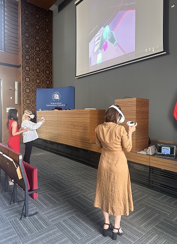 Trzy osoby stoją w auli KSSiP na rzutniku wyświetlana jest prezentacja. Dwie osoby mają założone gogle VR. 