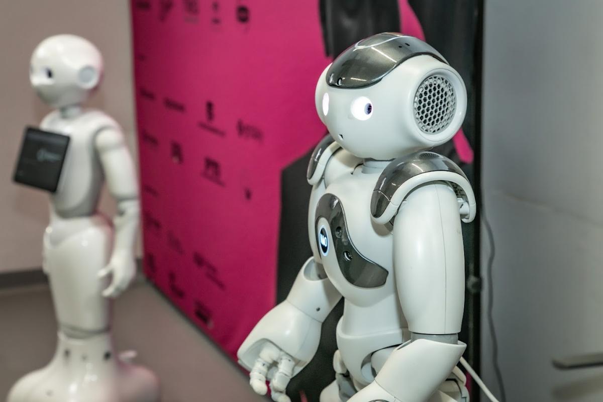 Dwa humanoidalne roboty stoją obok siebie w jasnym pomieszczeniu. Bliższy robot ma białą obudowę, duże okrągłe oczy i szare elementy na ramionach oraz głowie. Drugi robot jest biały i ma zamocowany z przodu tablet. W tle widać różowy baner z logotypami.