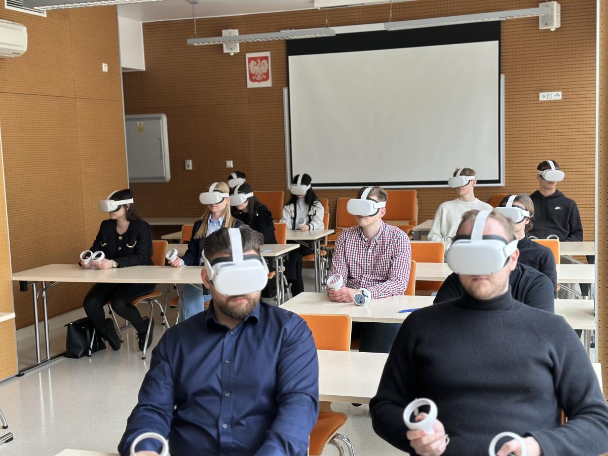 Grupa osób siedzących w nowoczesnej sali szkoleniowej, każdy z uczestników ma na głowie gogle VR i trzyma w rękach kontrolery. Osoby siedzą przy biurkach w rzędach, skupione na doświadczeniu wirtualnej rzeczywistości. W tle widać pusty ekran projekcyjny i godło Polski.