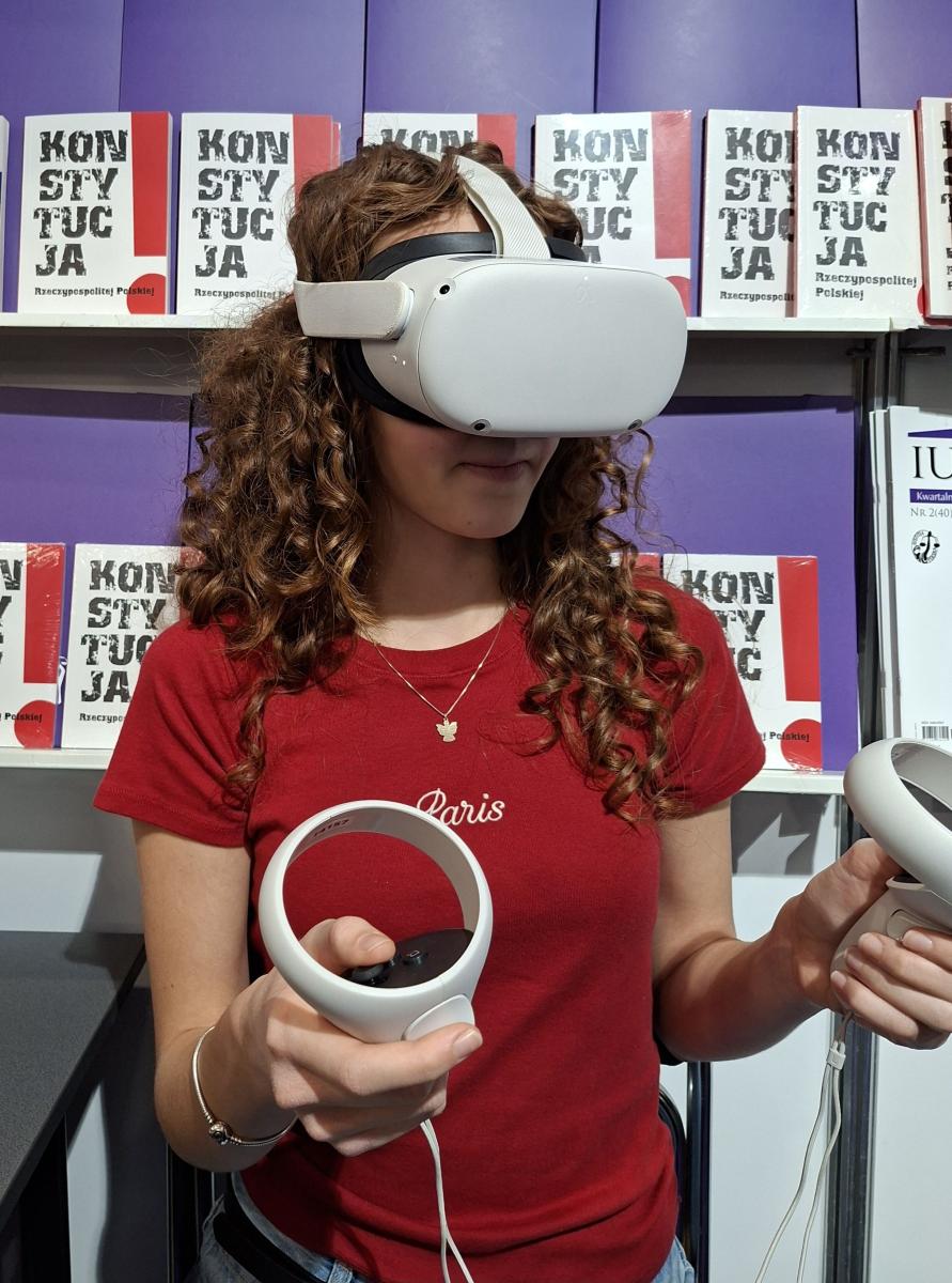 Młoda kobieta w czerwonej bluzce siedzi na krześle i korzysta z gogli wirtualnej rzeczywistości. W dłoniach trzyma kontrolery. Za nią widoczna jest ściana z półkami. Na półkach znajdują się książki z napisem Konstytucja Rzeczypospolitej Polskiej. 